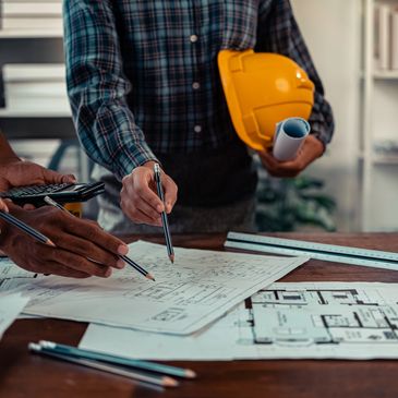 Engineers reviewing blueprints at a worktable with a yellow safety helmet.