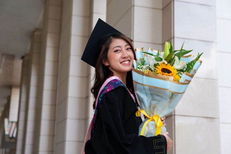 A young woman in her graduation gown and holding a bouquet of flowers, smiles confidently and celebrating the fruits of her hard work and dedication.
