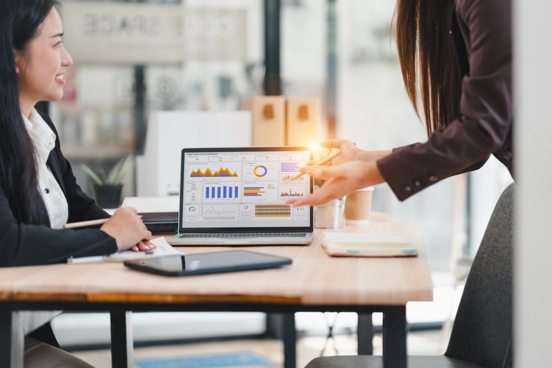 Businesswoman presenting data analysis on laptop screen to colleague in modern office setting, both women engaged in discussion with digital charts and graphs visible