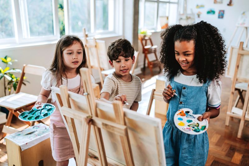Small group of diverse children, black and white students, gathered around canvas observing classmate painting during art lesson. Concept of childhood creativity, education, diversity, inclusion, teamwork and artistic expression in school environment.