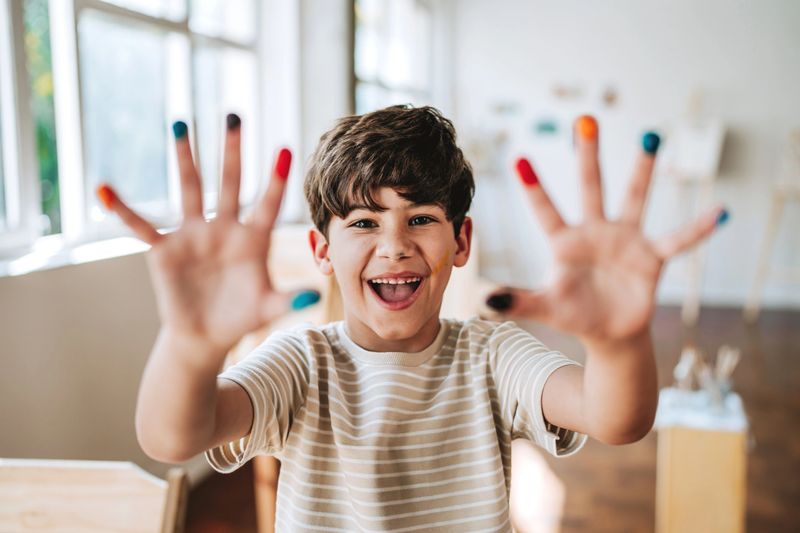Happy young boy looking at camera and smiling while showing hands covered with colorful paint. Child enjoying art class activity, concept of creativity, childhood, education and artistic expression.