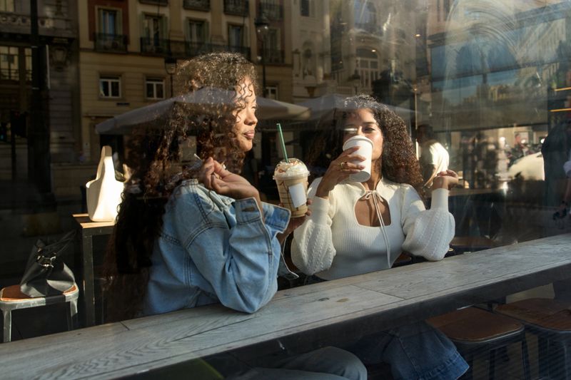 Two adult women sharing drinks and conversation inside a urban cafe, seen through the window with city reflections