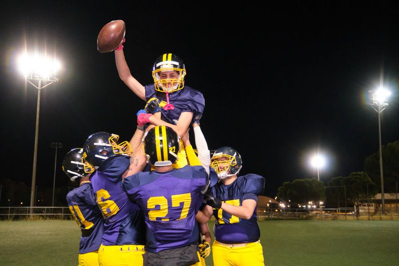 American football team lifting player holding ball, celebrating win on field at night