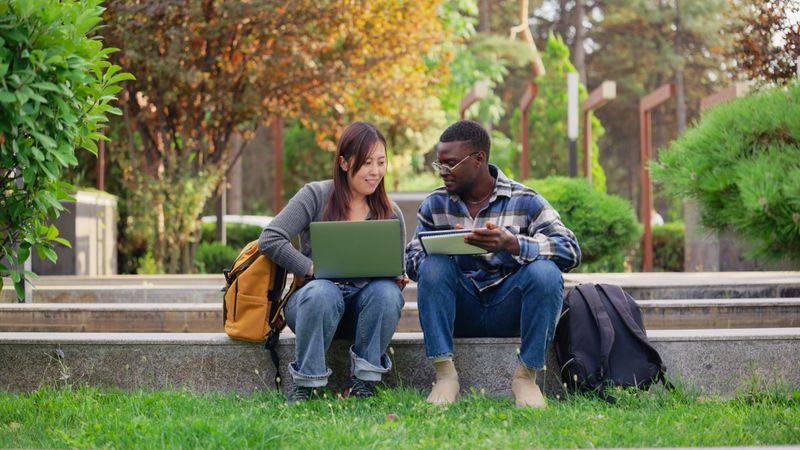 Two multiracial university students are sitting and studying together on a laptop at a campus.