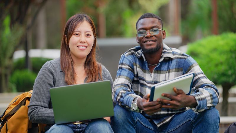 A Japanese female and a Black ethnic university students are sitting and looking at the camera with their laptop at campus.