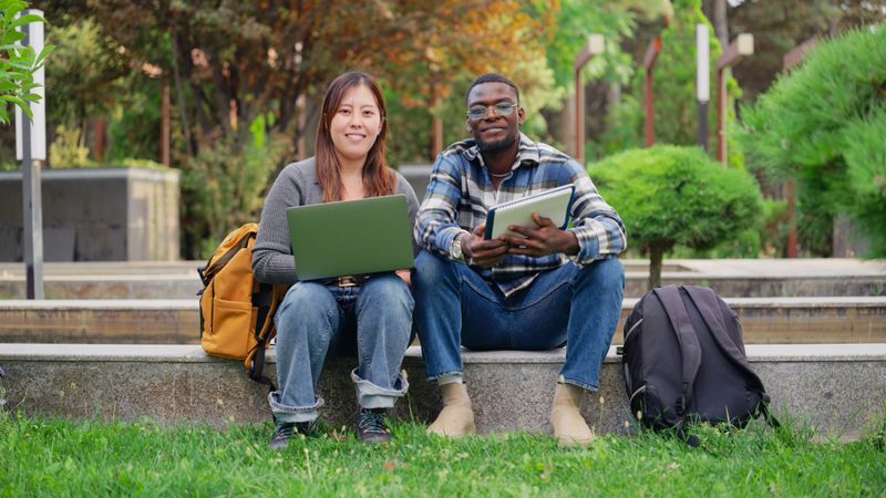 A Japanese female and a Black ethnic university students are sitting and looking at the camera with their laptop at campus.