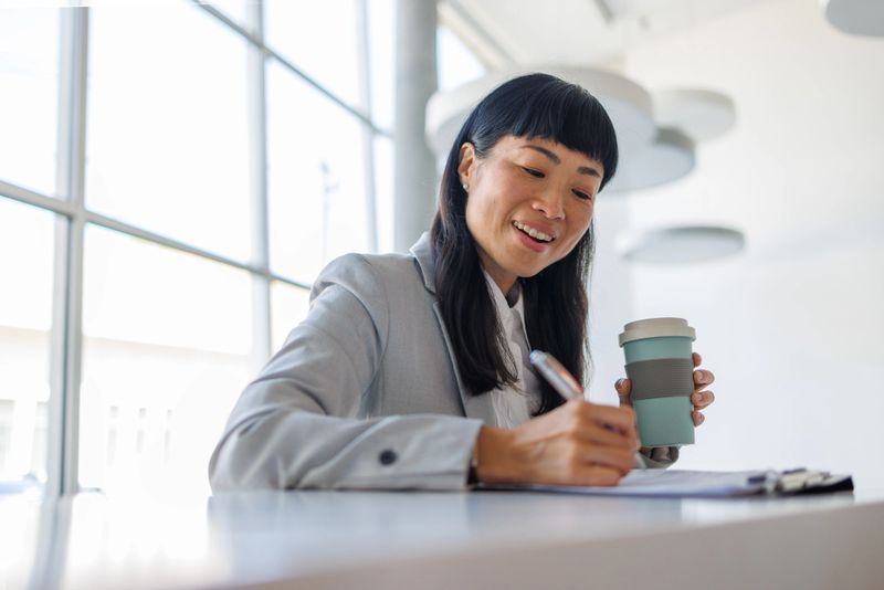 Smiling businesswoman writing on a document while holding a reusable coffee cup in a modern office