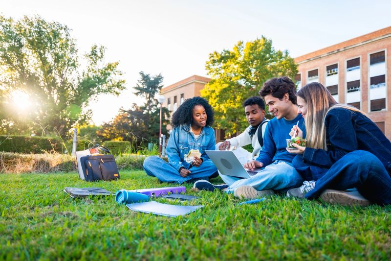 Group of four multicultural university students sitting on the campus lawn, sharing food and studying together around a laptop on a sunny day, smiling and collaborating outdoors