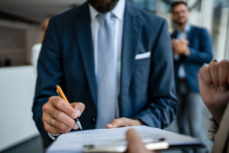 Mid adult businessman signing an important legal document, finalizing a business deal in a modern office, while another professional man watches in the background