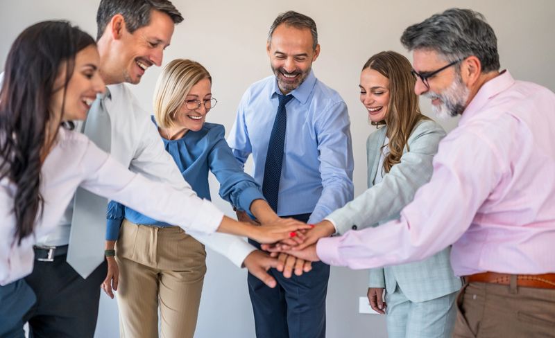 Diverse business team of six smiling colleagues stacking hands in an office, celebrating unity, teamwork and shared success