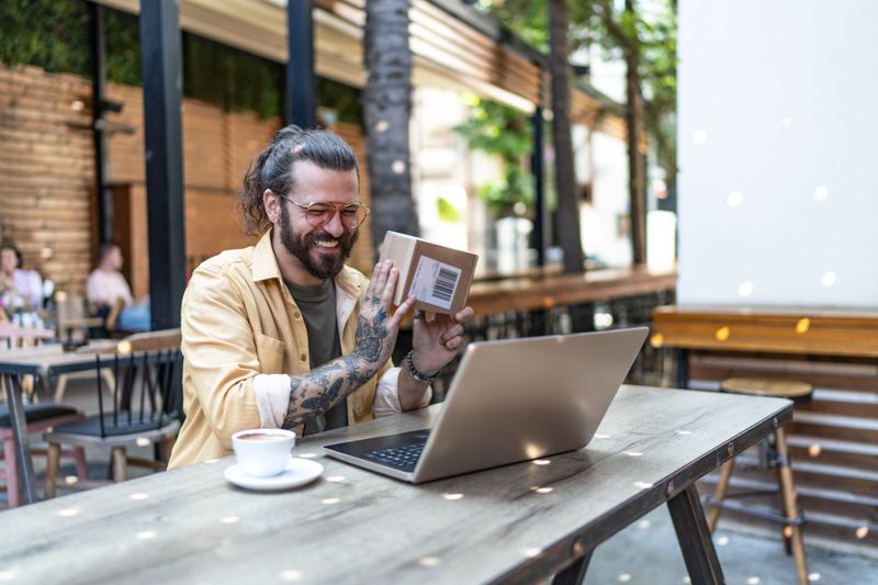 Smiling man with tattoos participating in a video call on his laptop from an outdoor cafe, holding up a small cardboard package, celebrating a successful online purchase