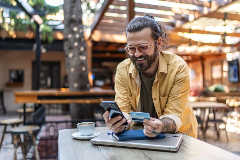Smiling mid-30s man using smartphone and credit card to complete a secure mobile payment at a cafe table with laptop and coffee, enjoying cashless convenience and online shopping convenience