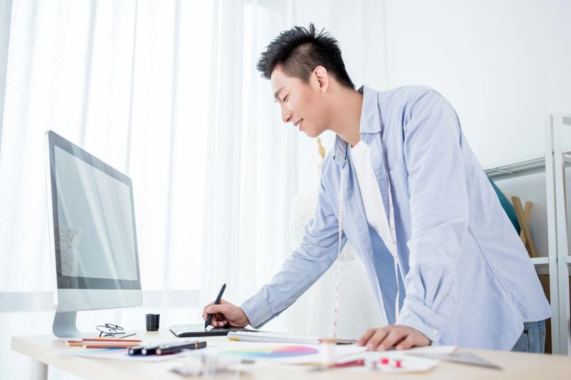 A young man is standing at a desk, working with a computer and various drawing tools. He is wearing a casual blue shirt and a white undershirt. The workspace is bright with natural light coming through the window. He appears to be focused on his task, using a pen and notebook while having a computer monitor in front of him.