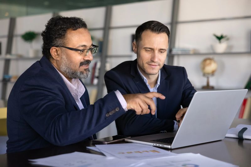 Two focused multinational businessmen working together at table, pointing at laptop screen, discussing or explaining strategy to client or colleague, analyzing financial charts, planning next steps