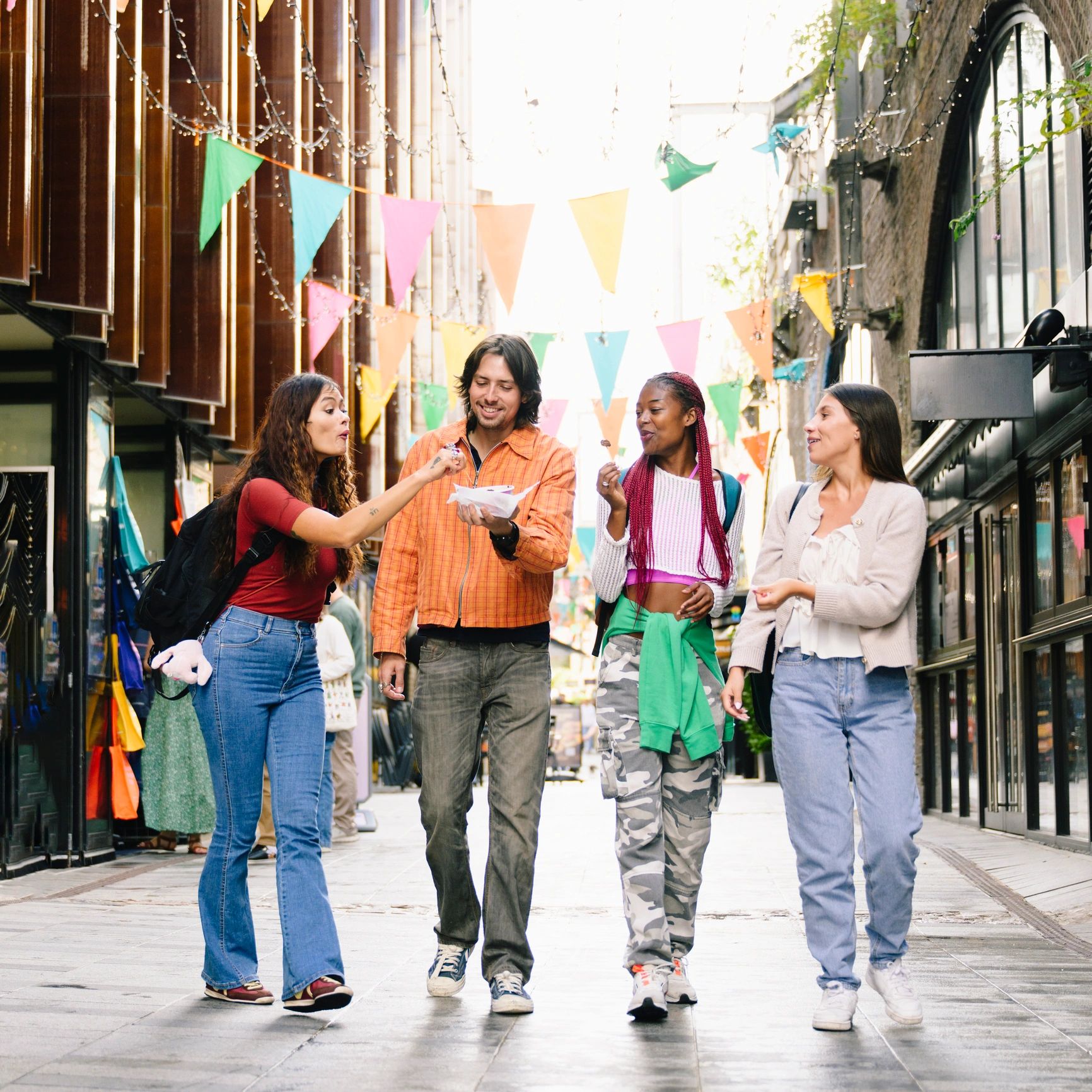 Four friends enjoy snacks and chat while walking through a festive street with colorful flags.
