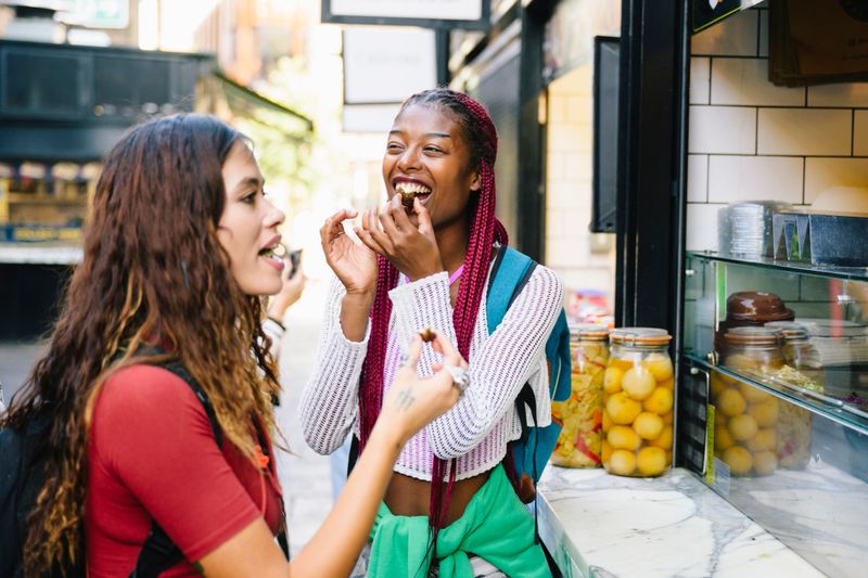 Two women eating food from takeaway counter, one laughing as she tastes street food