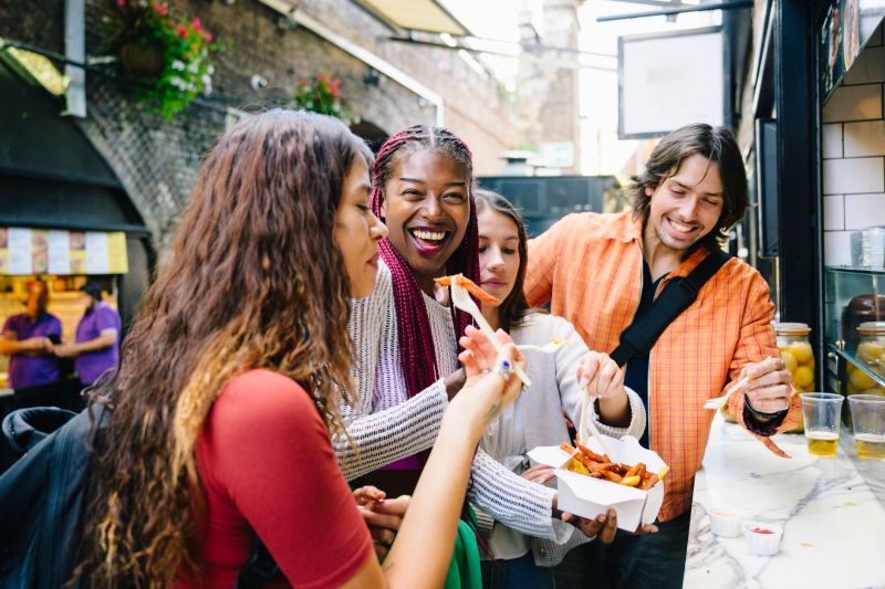 Woman in her 30s eating food from takeaway counter with fork, with three friends at the counter
