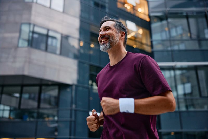 Mid-adult man jogging through a modern city street, smiling and looking up while exercising outdoors in casual sportswear, enjoying an active, healthy urban lifestyle
