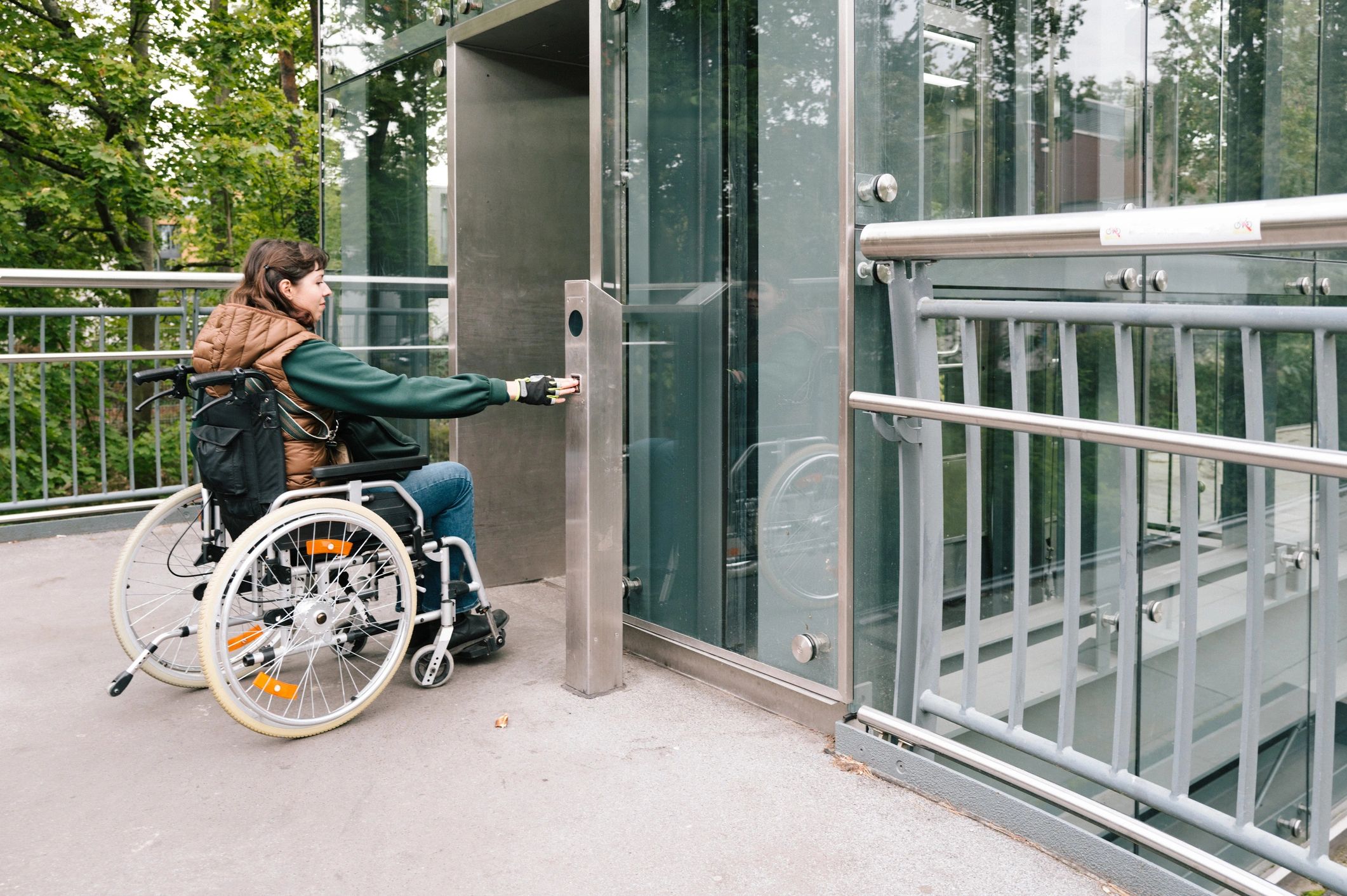 Woman in wheelchair pressing elevator button on an outdoor elevated path