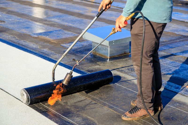 A roofer torches a membrane layer on a flat roof using a tiger torch to seal the asphalt bitumen, ensuring a durable waterproof layer.