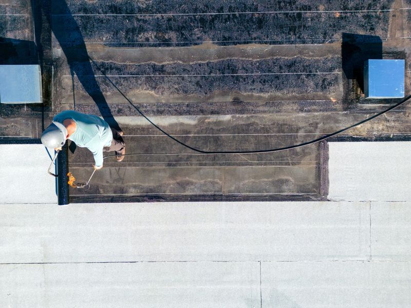 A roofer torches a membrane layer on a flat roof using a tiger torch to seal the asphalt bitumen, ensuring a durable waterproof layer.