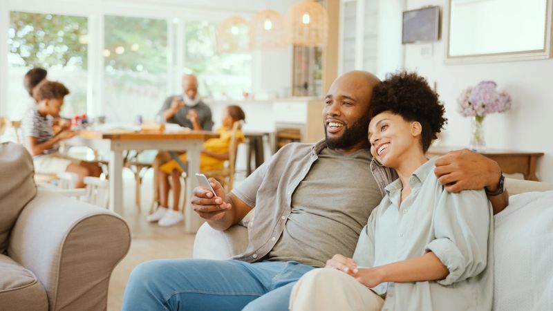 Couple At Home On Sofa Streaming Show Or Movie To TV With Multi-Generation Family In Background