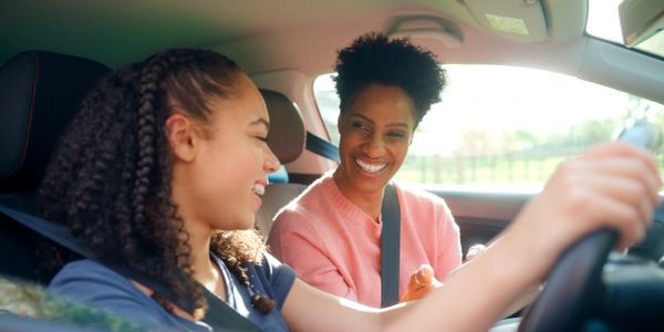 A smiling woman teaches a teenage girl to drive inside a car.