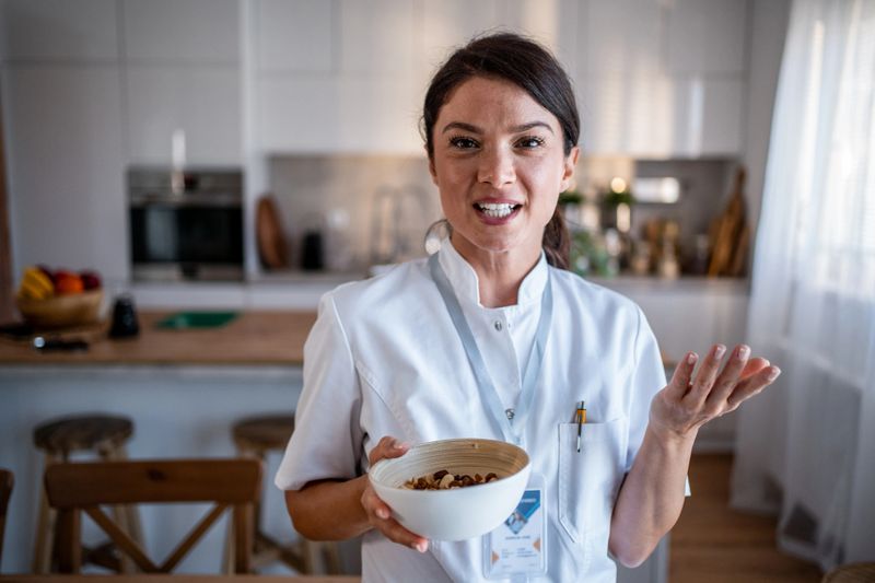 Female nutritionist in a modern kitchen holding a bowl of nuts, smiling and explaining healthy eating and plant-based snack options during an in-person or remote consultation