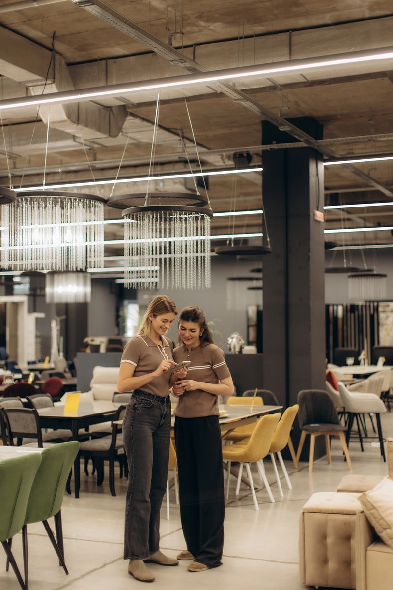 Two women discussing a purchase while shopping in a contemporary furniture store featuring sleek chairs, chandeliers, and a modern industrial design. The environment showcases stylish and functional home dÃ©cor pieces.