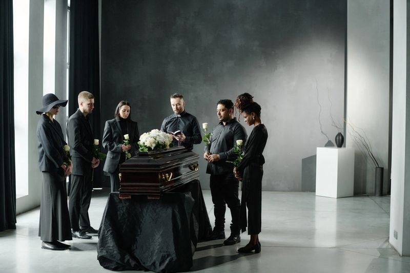 Group of middle aged and young adult men and women standing around closed casket holding roses during funeral ceremony in modern room