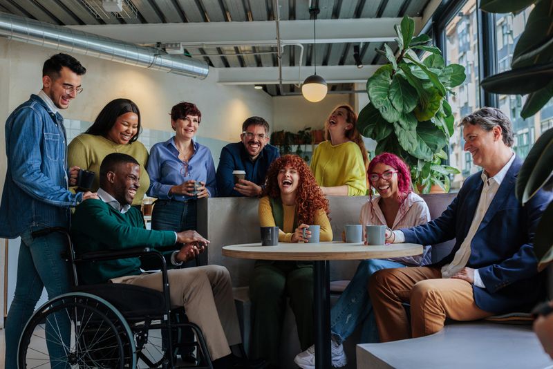 Happy multiracial colleagues sharing a coffee break, laughing and bonding in a wheelchair accessible, inclusive workplace