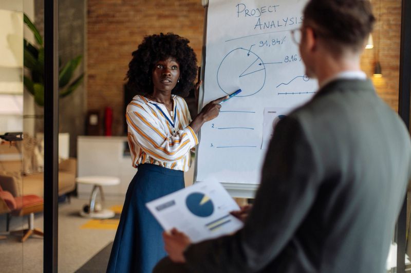 Professional woman standing by a flip chart explaining project data insights to her coworker during a collaborative brainstorming session in a contemporary workspace