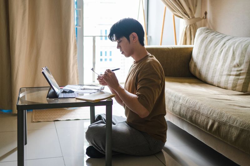 A young Asian man sits on the living room floor with a laptop and calculator, seriously calculating finances and looking at bills.