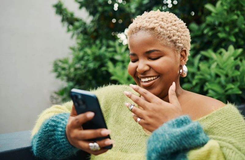 A joyful Black woman with short blonde hair and a warm smile enjoys a phone call while sitting outdoors, surrounded by lush greenery.