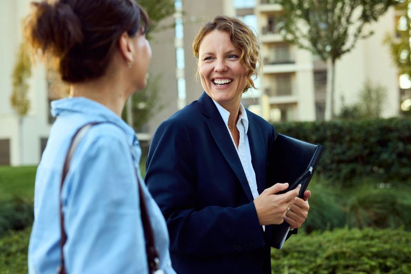 Two women in professional attire converse outdoors near a modern building, smiling while exchanging documents.
