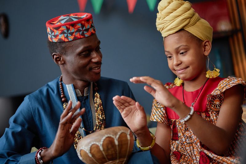 Black man teaching Black girl drumming on traditional drum during Kwanzaa celebration, both wearing cultural attire, girl smiling and focusing on learning rhythm