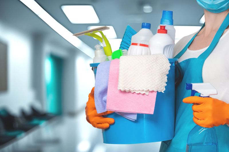 Cleaning supplies prepare for a deep clean in a hospital corridor. A person in protective gear holds a bucket full of cleaning products in a hospital corridor.