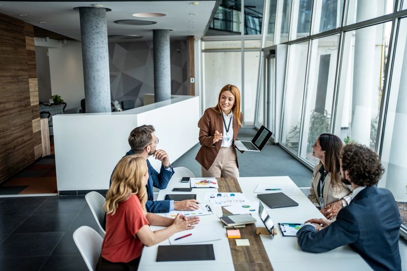 Businesswoman standing and explaining financial results to colleagues, showing charts and graphs on a laptop screen to a group of five professionals sitting at a conference table