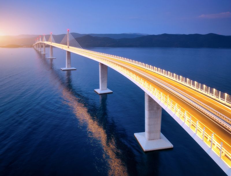 Aerial view of modern Peljesac bridge and blue sea at sunset. Croatia. Bridge with illumination, mountains, reflection and purple sky at sunset. Top drone panoramic view of road, street lights at dusk