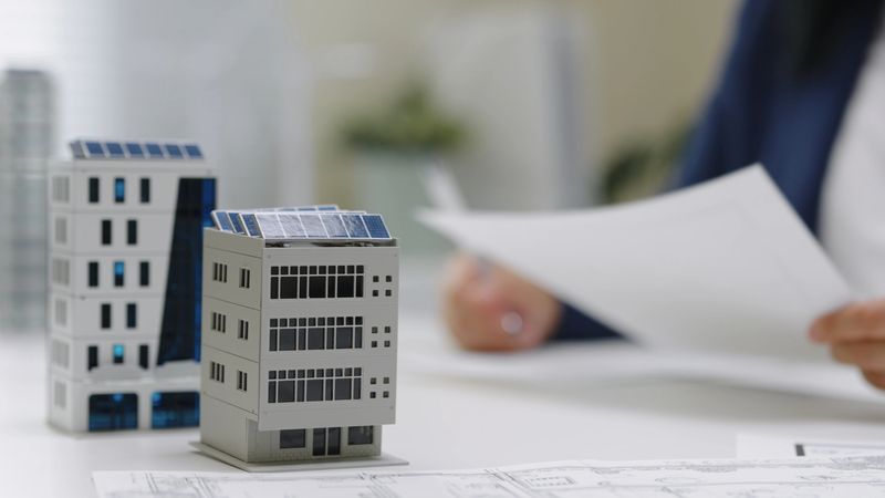 Businesswoman architect reviewing with construction architecture blueprint with office building exterior and 3D model with solar panels planning or design meeting on sustainable urban development.