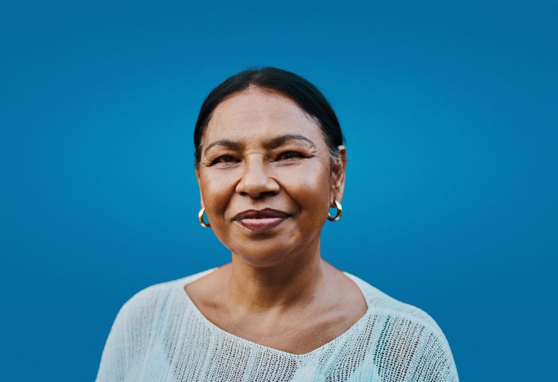 Portrait of a confident middle aged Latina woman wearing a white top and gold hoop earrings smiling gently with a clear blue background evoking an authentic and warm expression.