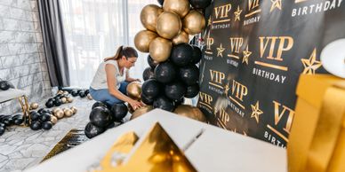 Woman arranging black and gold balloons for a VIP birthday party backdrop.