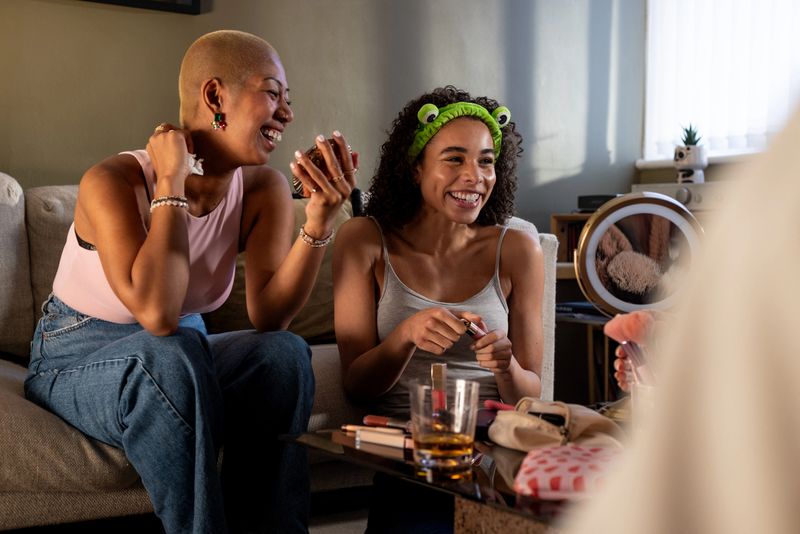 A medium shot of two cheerful women sitting together in a cosy living room, applying makeup and smiling as they share beauty products. A tabletop in front of them is filled with cosmetics, brushes, and drinks, creating a lively and authentic lifestyle scene. They are all preparing and getting ready for their graduation.Videos are available similar to this scenario.