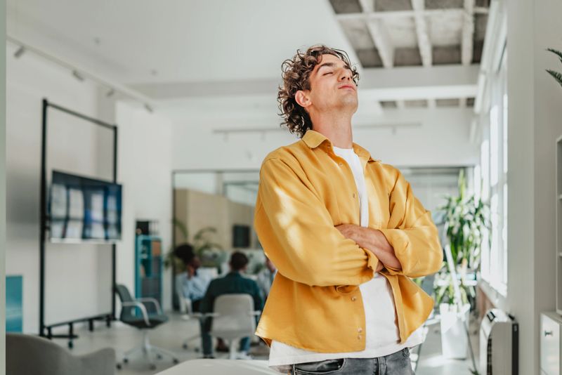 A young professional stands confidently in a contemporary office space, taking a moment to reflect while surrounded by coworkers engaged in a meeting. The atmosphere promotes teamwork and connection.