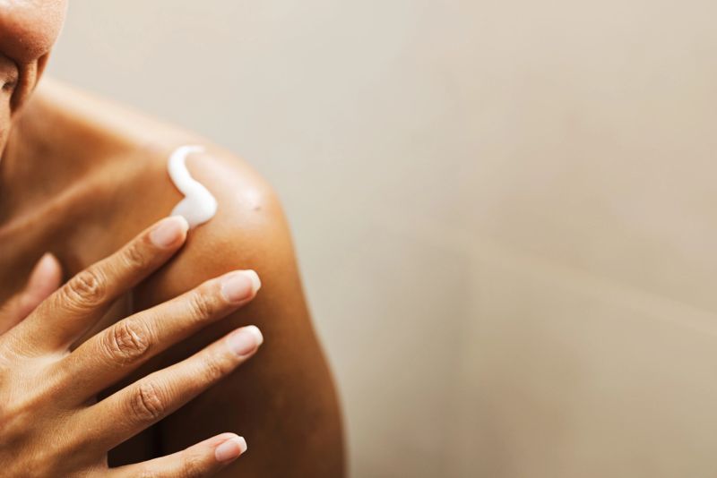 A close-up shot of a woman applying lotion to her shoulder, highlighting skincare, self-care, and wellness with a calm, intimate moment focused on skin health and moisturizer.