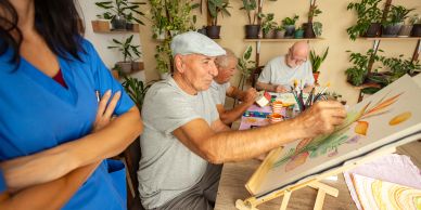 Elderly men painting in a cozy, plant-filled room with a caregiver.