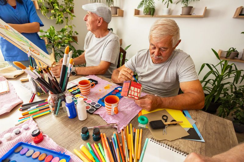A cheerful caregiver stands beside an elderly man drawing on an easel, highlighting supportive art therapy for seniors in a sunny, plant-filled environment.