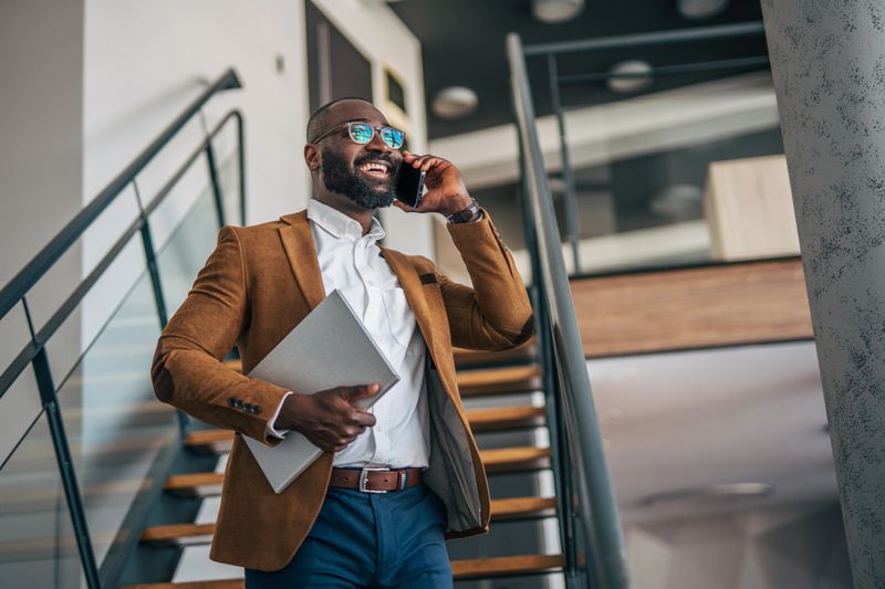 Happy young adult black man in a modern office building making a business phone call while confidently walking up a contemporary staircase, holding a laptop
