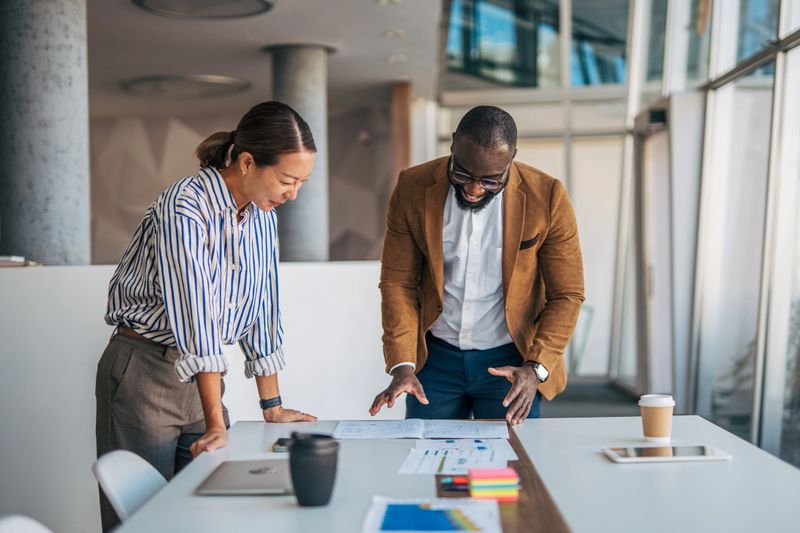 Multiracial business partners analyzing reports and charts on a meeting table, discussing strategies and planning new business goals during a collaborative session in a bright office environment