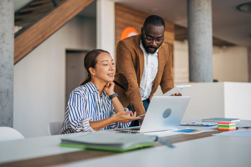 Diverse business colleagues collaborating over laptop and tablet in a bright modern office, reviewing data and planning a project together with focused, friendly teamwork and tech tools